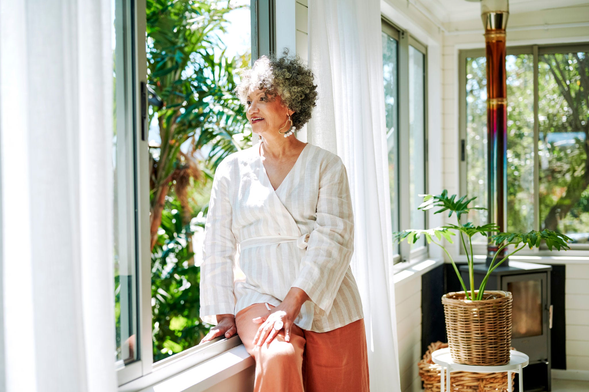Smiling mature woman sitting on a window sill and looking out at the view while relaxing at home