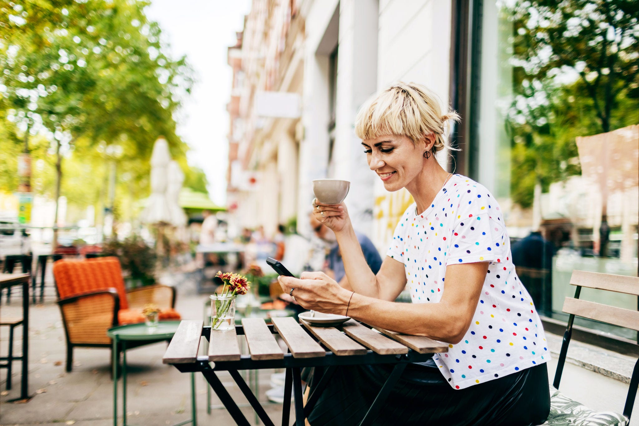 A woman sitting at a table outside a cafe looking at her smartphone while drinking a cup of coffee.